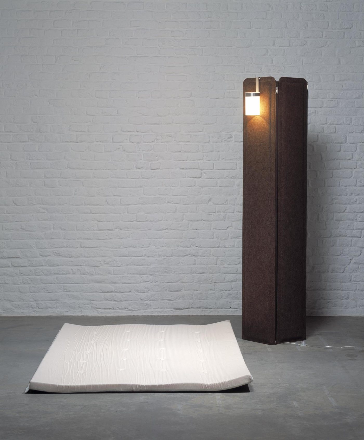 Jim de matali crasset : la colonne de feutre brun et sa lampe, avec le matelas posé au sol