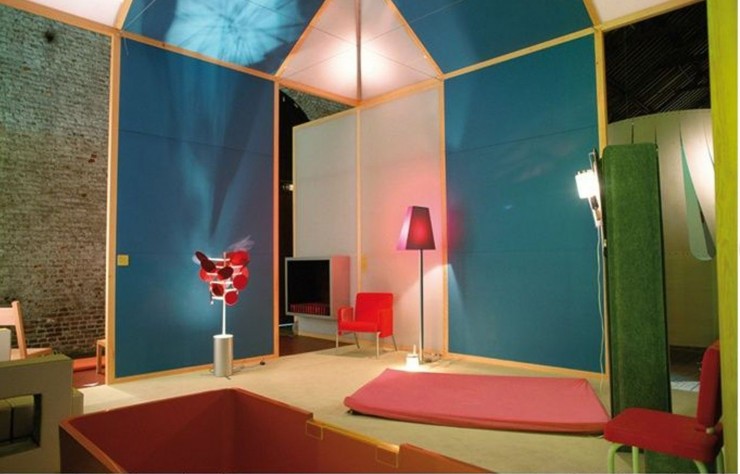Une installation aux parois bleues et blanches, avec un tapis rose au sol, une lampe rouge et, à droite, une colonne de feutre vert